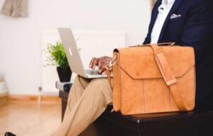 person-sitting-on-a-black-chair-with-a-briefcase-next-to-them-typing-on-a-laptop